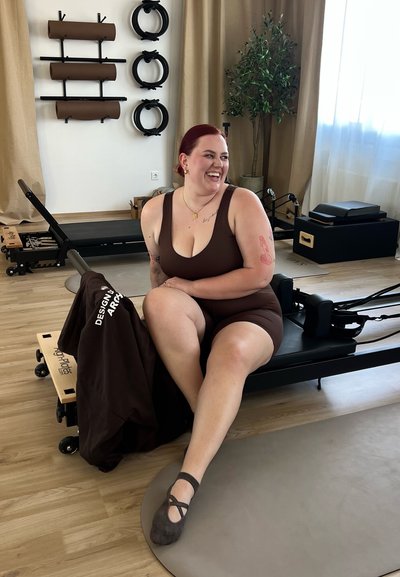 Reformer de pilates marrón con detalles en negro, esterilla gris, plataforma de madera con ruedas y una persona vestida con un atuendo atlético marrón sentada en él.