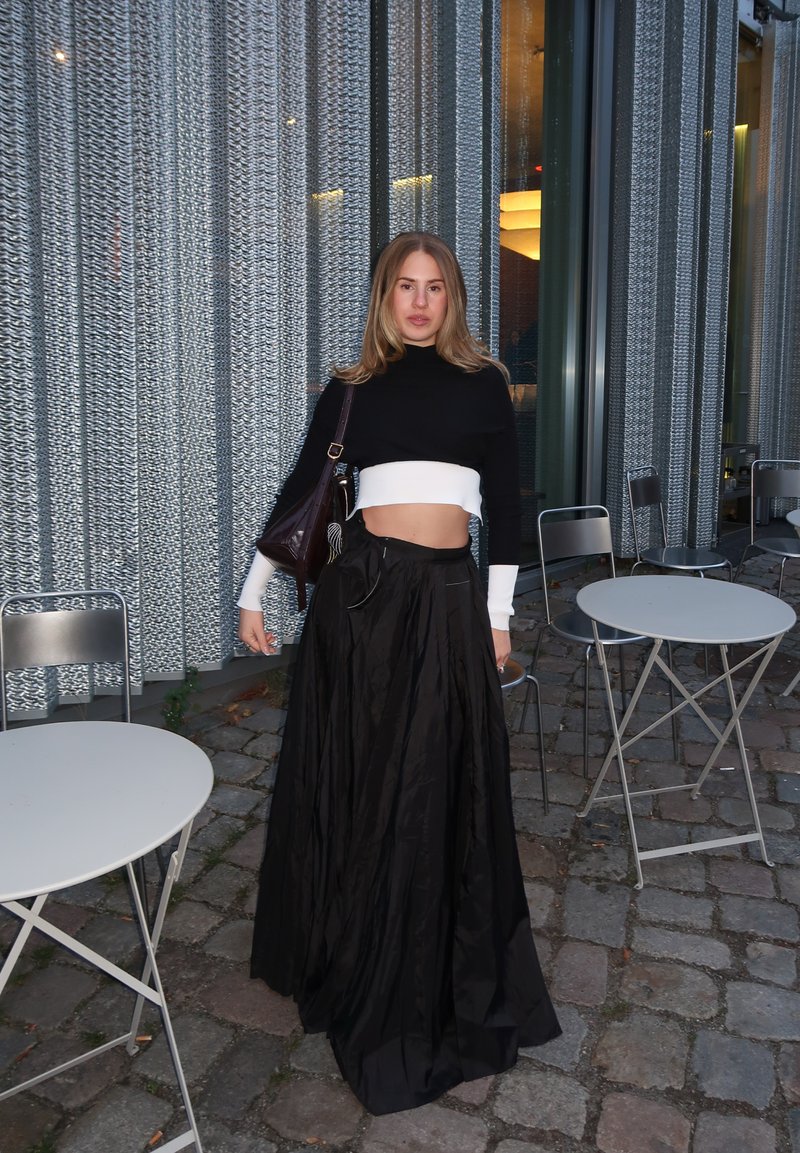 Gonna maxi nera realizzata in tessuto fluido abbinata a un maglione corto bianco e nero. Una borsa marrone e un'atmosfera da caffè all'aperto.