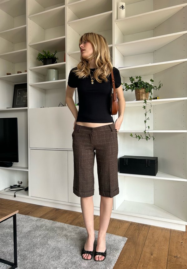 Svart åtsittande topp, bruna rutiga capribyxor och svarta platta sandaler. Naturligt hår och halsband; inomhusmiljö med hyllor och växter.