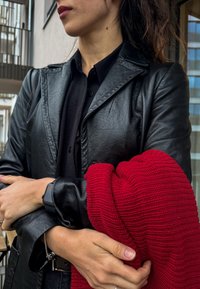 Μαύρο δερμάτινο μπουφάν, μαύρο πουκάμισο από κάτω, κρατώντας ένα κόκκινο πλεκτό πουλόβερ. Τα χαρακτηριστικά περιλαμβάνουν ομαλή υφή, εφαρμοστή σχεδίαση και μαλακό ύφασμα.