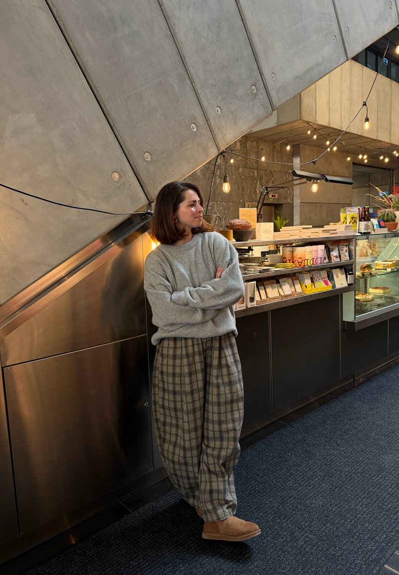 Kvinna i grå tröja och rutiga byxor står med armarna i kors inne i en butik nära en monter med förpackade varor och desserter.