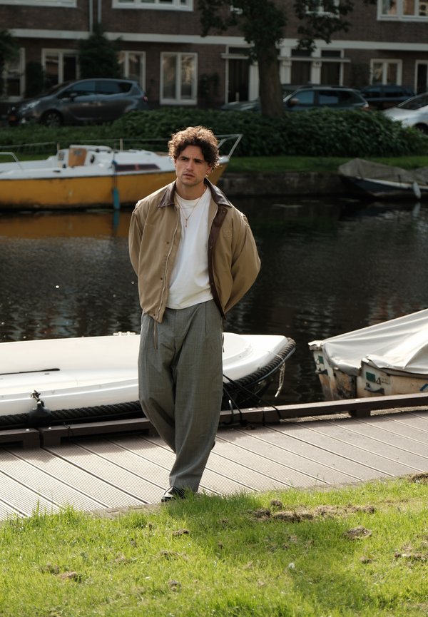 Junger Mann mit lockigem Haar steht an einem Steg in der Nähe von Booten, trägt eine beige Jacke, weißes Hemd, graue Hose an der Uferpromenade mit parkenden Autos und Gebäuden im Hintergrund.