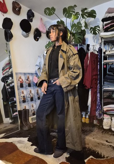 Persona con un abrigo largo beige, camisa negra y vaqueros oscuros, de pie en una habitación con sombreros, ropa, botas y plantas verdes.