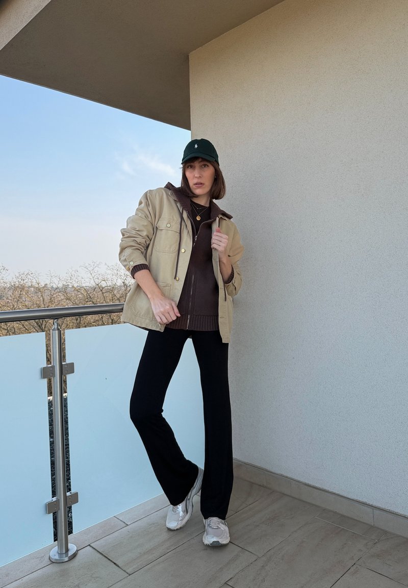 Vrouw draagt een beige jas, donkervest met capuchon, zwarte broek, witte sneakers en een groene pet, staand op een balkon met reling en een heldere lucht op de achtergrond.