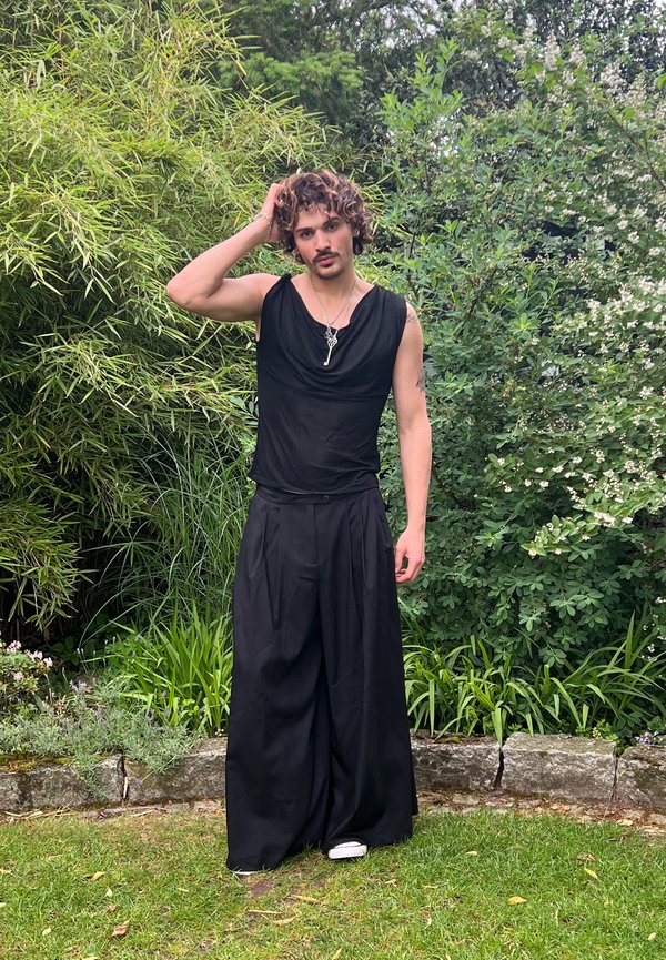 Persona con cabello rizado que lleva una camiseta negra sin mangas y pantalones anchos negros, de pie sobre el césped, tocándose la cabeza, con plantas verdes y un borde de piedra detrás.