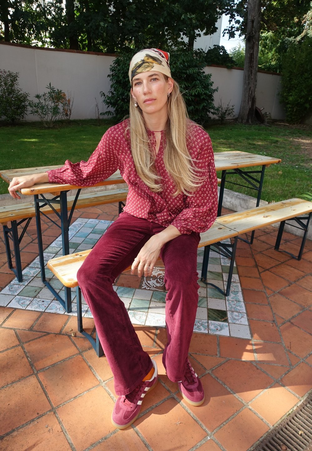 Bordeauxfarbene Cord-Hosen mit Schlag, gemusterte burgunderfarbene Bluse und burgunderfarbene Sneakers mit weißen Streifen. Model sitzt auf einer Holzbank.