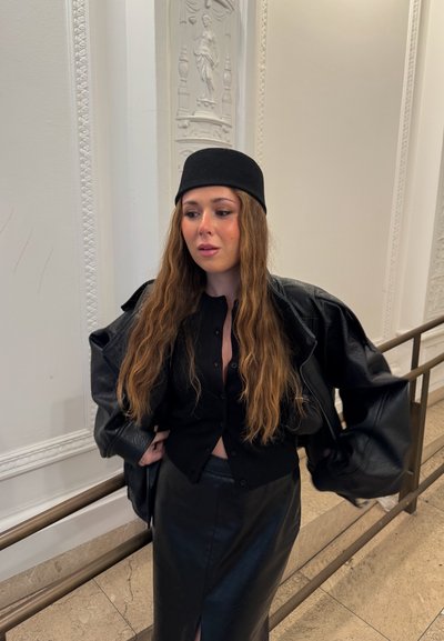 Mujer con cabello largo y ondulado que lleva un sombrero negro, blusa negra con botones, chaqueta de cuero negra y falda de cuero negra, de pie en el interior junto a una barandilla.