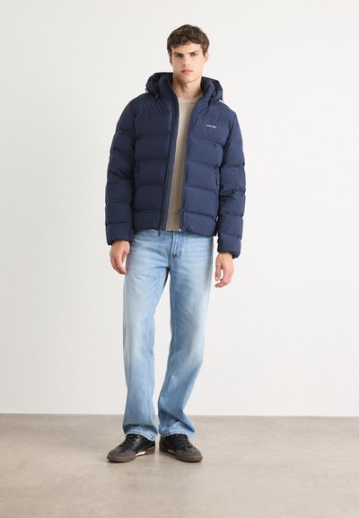 Chaqueta acolchada navy con capucha, que presenta un cierre de cremallera y un diseño acolchado. Lleva puesta sobre una camisa beige y combinada con jeans de color azul claro y zapatillas negras.