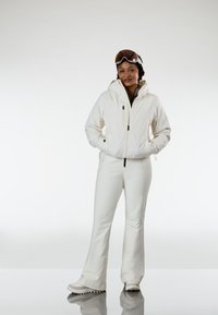 Άτομο ντυμένο με λευκή στολή σκι, φοράει γυαλιά προστασίας στο κεφάλι, στεκεται με τα χέρια στις τσέπες του σακακιού μπροστά σε απλό φόντο.