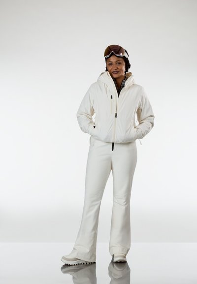 Persona vestida con un traje de esquí blanco, con gafas en la cabeza, de pie con las manos en los bolsillos de la chaqueta contra un fondo liso.