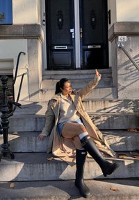 Zwarte rubberen regenlaarzen met witte accenten en gesp, gecombineerd met een beige trenchcoat en een lichte trui, zittend op stenen trappen.