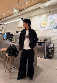 Jeune homme en veste noire et pantalon ample, debout près du comptoir d'un café avec des tabourets, des sacs et des panneaux de menu sur un mur en briques blanches.