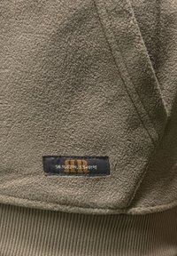 Material fleece verde măsliniu cu suprafață texturată, având o etichetă neagră cu litere aurii și coordonate geografice.