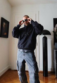 Felpa nera con tasca frontale, pantaloni cargo grigi con tasche laterali e occhiali da sole neri. L'ambiente presenta un arredamento moderno e opere d'arte.