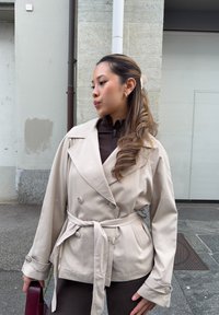Beige dobbeltradet trenchcoat med bindebælte, med brede revers og knapdetaljer, båret over en brun top, med en håndtaske i hånden.