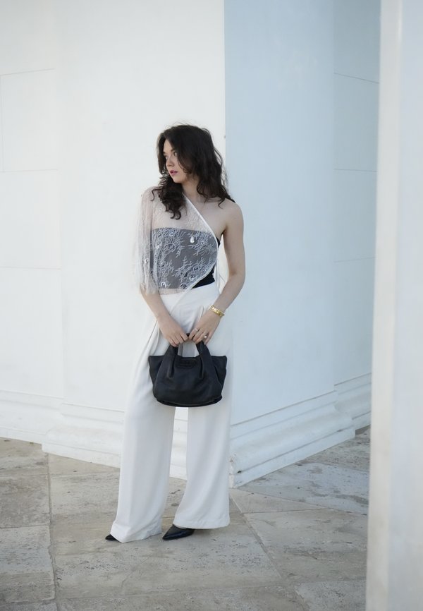 Frau in weissen weiten Hosen und einem asymmetrischen Spitzenoberteil, die eine schwarze Handtasche hält und vor einem weißen architektonischen Hintergrund steht.