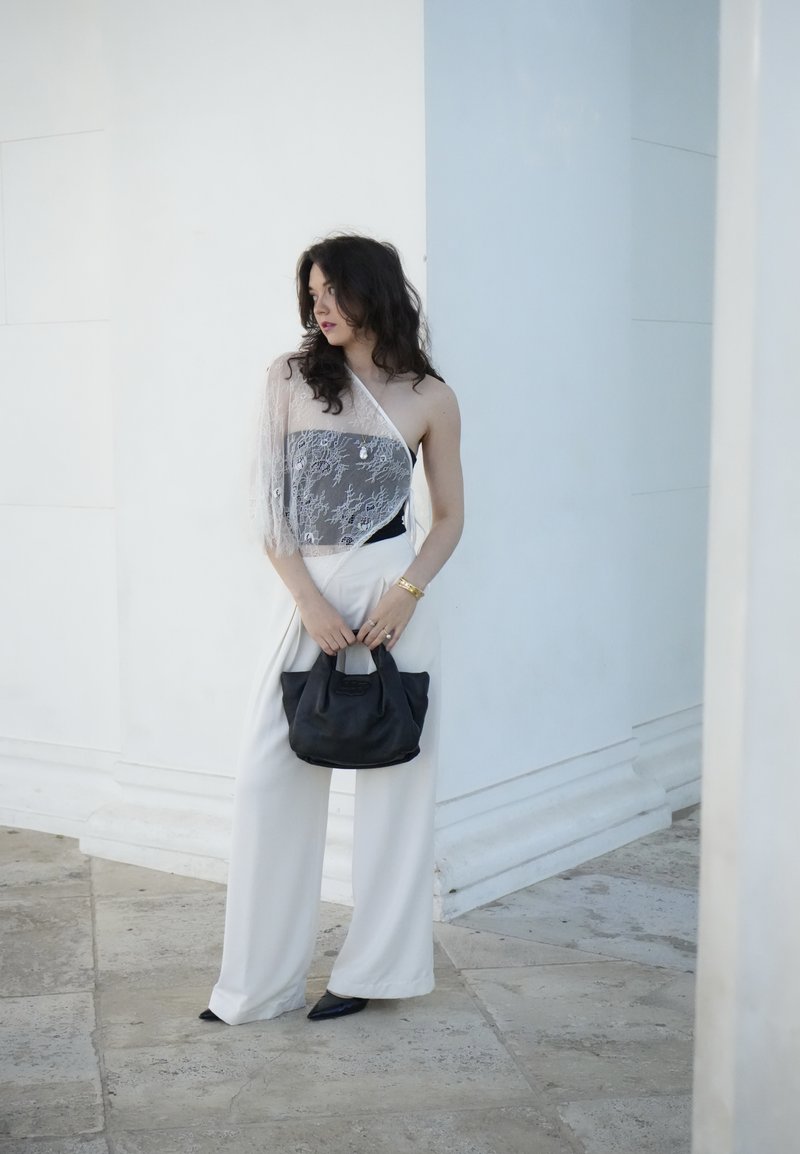 Femme en pantalon blanc large et top en dentelle à une épaule tenant un sac à main noir, debout devant un fond architectural blanc.