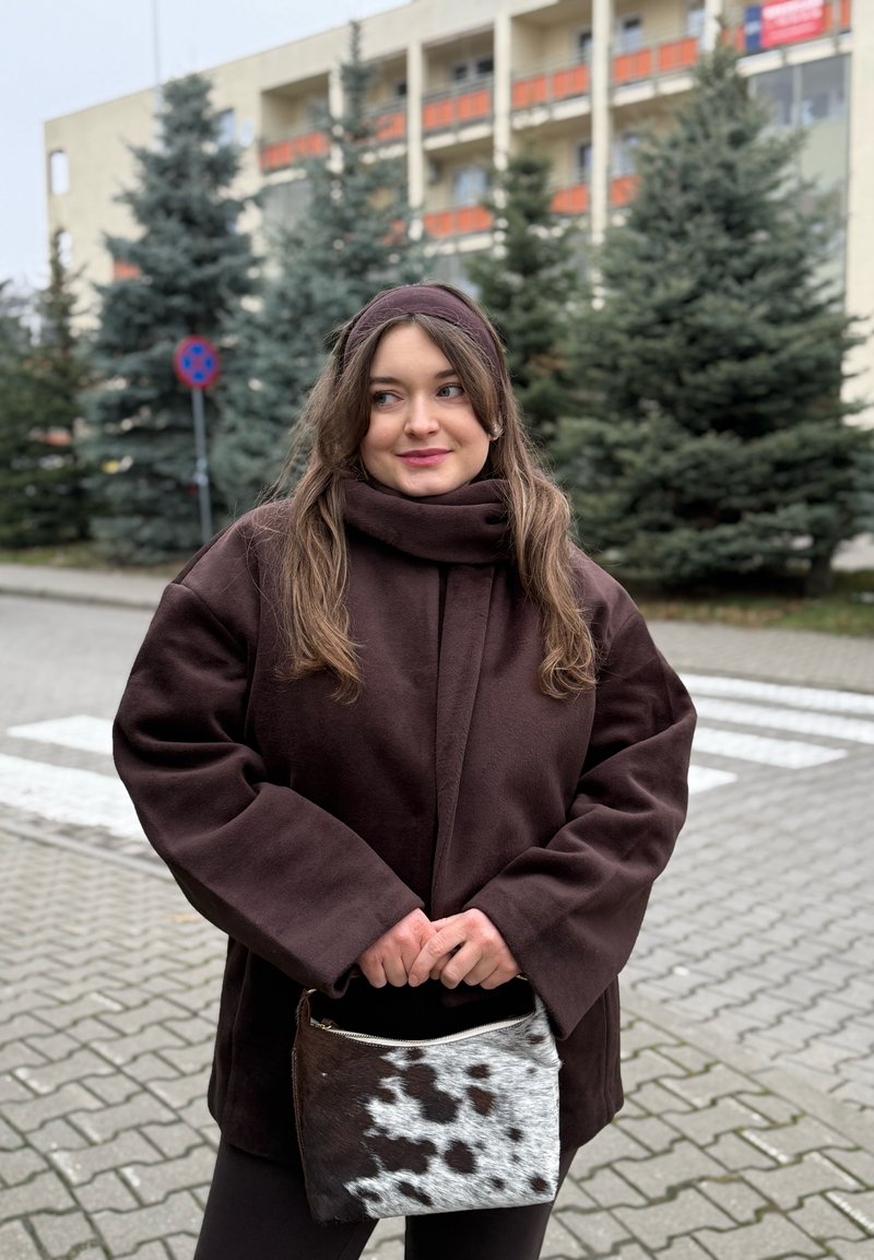 Veste polaire marron à manches larges et col montant, associée à un sac à main texturé noir et blanc à pois. Arrière-plan urbain avec des arbres.