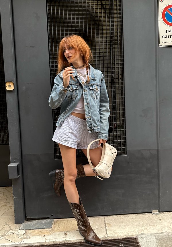 Jeansjack over een witte crop top, gestreepte shorts, bruine geborduurde cowboylaarzen en een witte handtas met een schouderband, tegen een grijze muur.