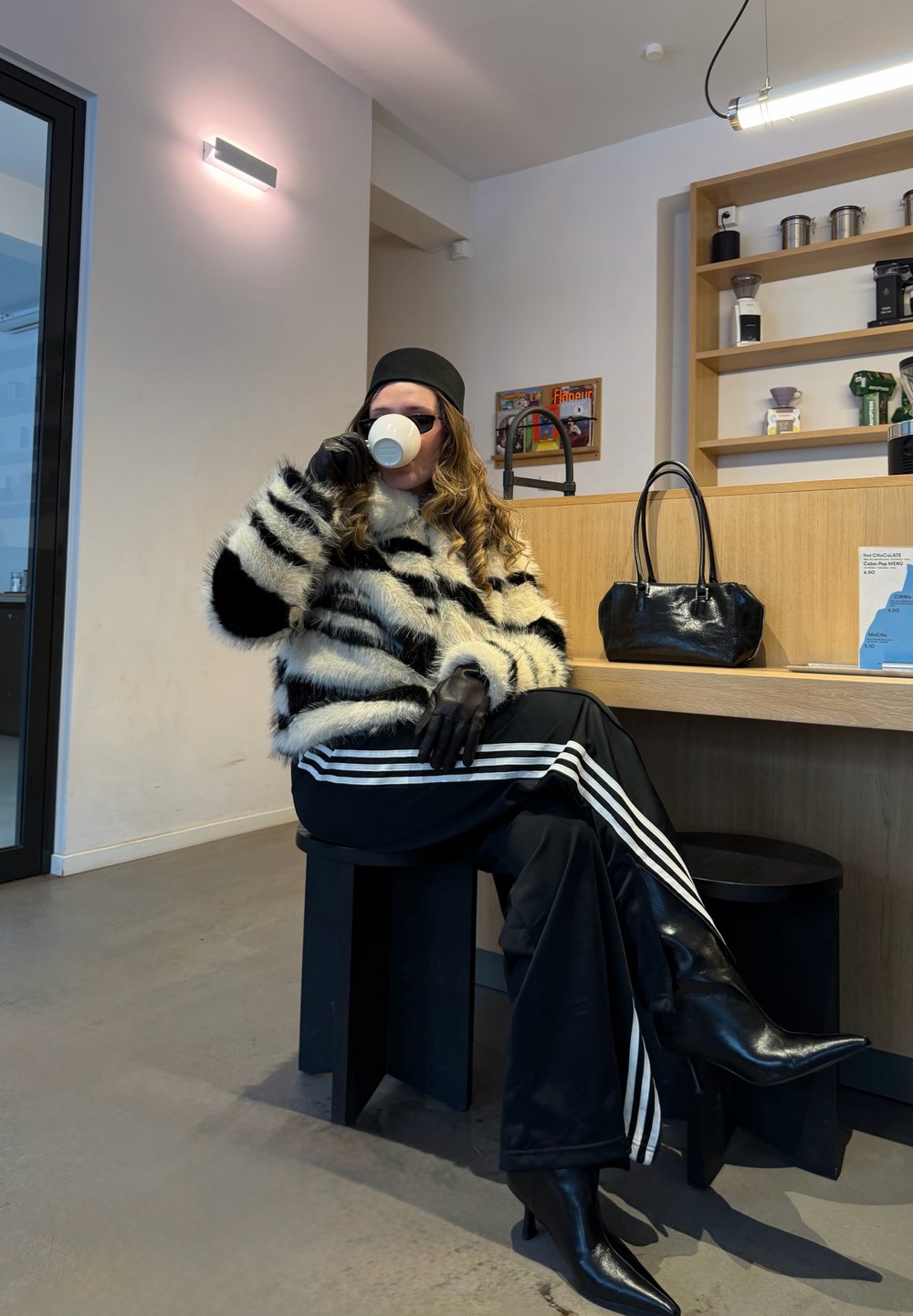 Personne portant un manteau en fausse fourrure rayé, des gants en cuir noir et des bottes, assise sur un tabouret, buvant dans une tasse blanche dans un cadre de café.