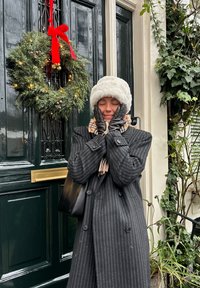 Schwarz gestreifter Mantel, weißer Pelzhut und schwarze Handschuhe, stehen neben einer dunkelgrünen Tür, die mit einem Weihnachtskranz und rotem Band geschmückt ist.