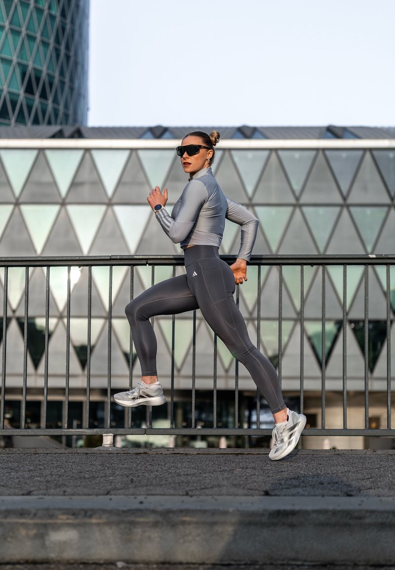 Femme en tenue de sport grise et lunettes de soleil courant à l'extérieur près d'un bâtiment en verre géométrique, en plein mouvement avec une rampe au premier plan.