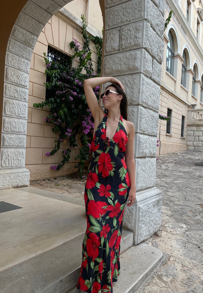 Maxi φόρεμα με σχέδιο halter neck, μαύρο ύφασμα με ζωντανά κόκκινα λουλουδάτα μοτίβα και ρο-flowing σιλουέτα, μπροστά σε μια πέτρινη αψίδα.