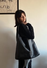 Grand sac hobo en cuir noir avec une texture lisse, une large bandoulière et une forme décontractée. Présente un design minimaliste sans quincaillerie visible.