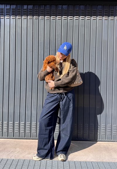 Una persona con un suéter marrón sostiene a un pequeño perro marrón de pelo rizado. Lleva unos pantalones de mezclilla oscuros y holgados, y una gorra de béisbol azul, de pie contra una pared gris.