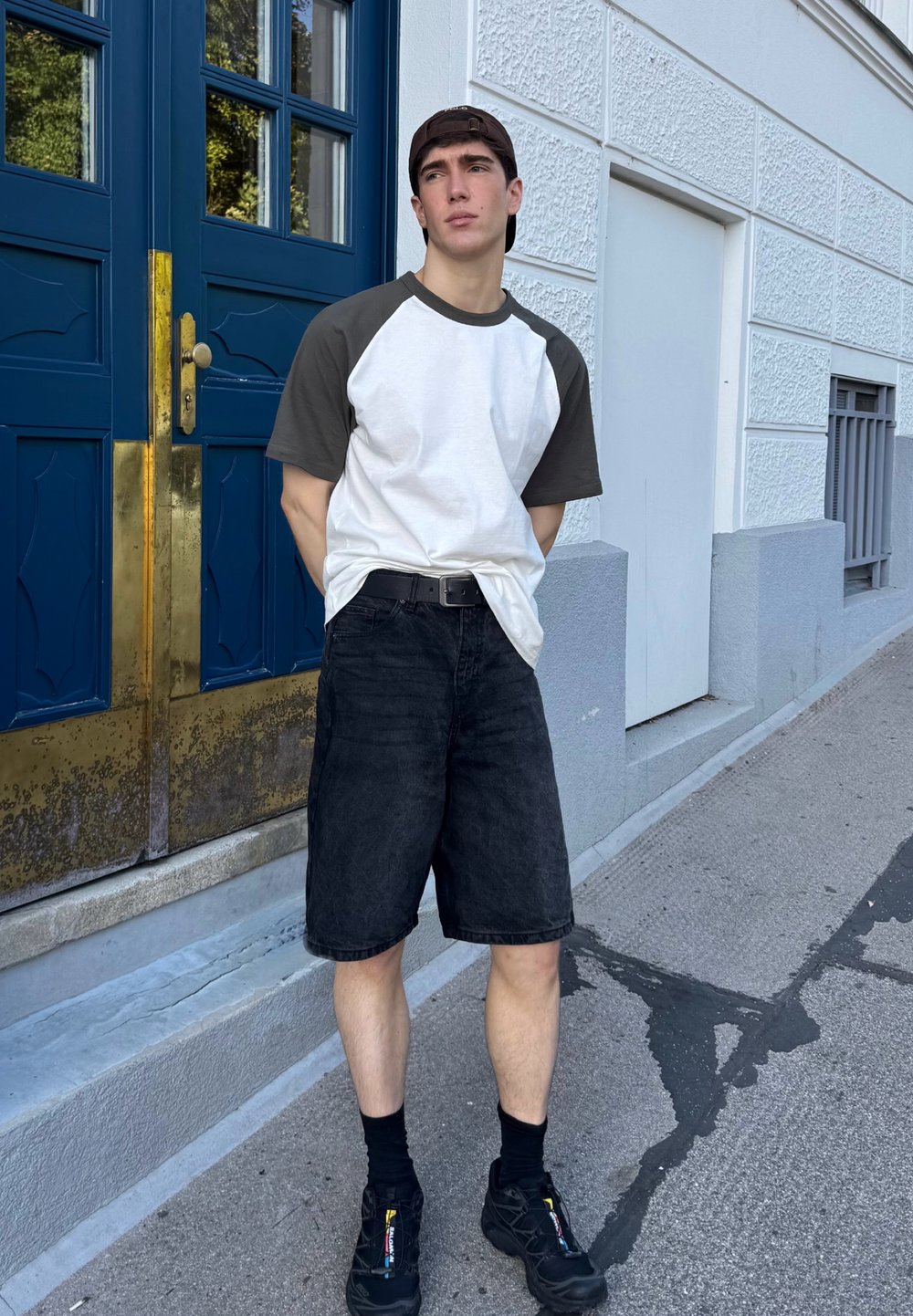 Junger Erwachsener in einem weißen und dunkelgrauen Kurzarm-Raglan-Shirt, schwarzen Denim-Shorts, schwarzen Turnschuhen und einer dunklen Mütze, der neben einer blauen Tür steht.