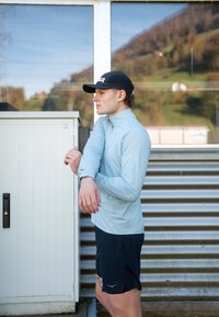 Gaiši zila garroku sporta krekls ar augstu apkakli un tumši šorti. Aprīkots ar atstarojošiem akcentiem. Modele pieskaras piedurknei pie balta skapja.