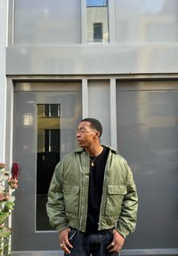 Giacca bomber imbottita verde con zip frontale, due tasche, polsini a costine, indossata sopra una maglietta nera. Sullo sfondo si vedono una porta grigia e piante.