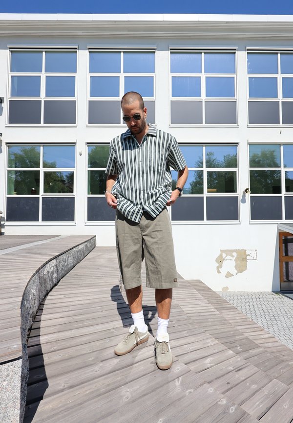 Camisa de mangas curtas às riscas em verde e branco, calções cinza claro, sapatilhas cinza e meias brancas, de pé num caminho de madeira perto de um edifício.