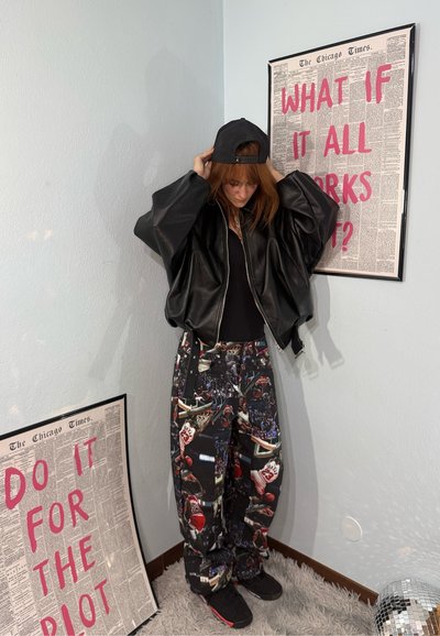 Persona ajustándose una gorra negra, con una chaqueta de cuero negra extragrande y pantalones estampados, de pie entre dos carteles enmarcados con texto rojo intenso.