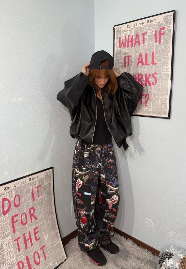 Personne ajustant une casquette noire, portant une veste en cuir noire oversize et un pantalon à motifs, debout entre deux affiches encadrées avec du texte rouge vif.