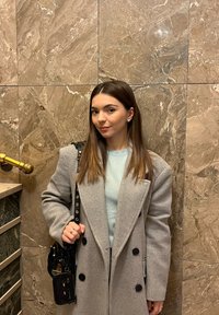 Lysegrå dobbeltradet frakke over en lys blå sweater, der holder en lille sort håndtaske med kædestrop. Marmorvæg i baggrunden.