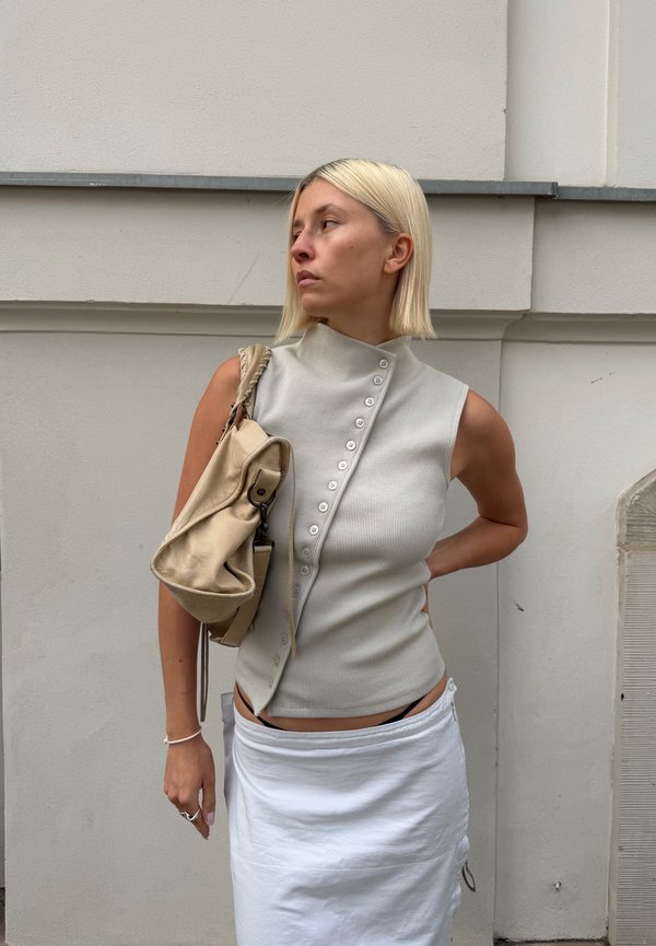 Blonde Frau trägt ein ärmelloses beiges Knopftop und weißen Rock, hält eine beige Tasche über der Schulter, steht vor einer hellen Wand.