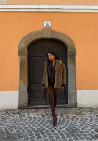 Manteau en fausse fourrure marron avec des détails texturés, associé à des collants imprimé léopard. Accessoirisé d'un sac à main rouge, debout près d'une porte en bois.