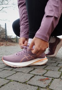 Zapatillas deportivas moradas con detalles en naranja, diseño ligero, parte superior texturizada y cordones prominentes, combinadas con leggings negros y una blusa malva.