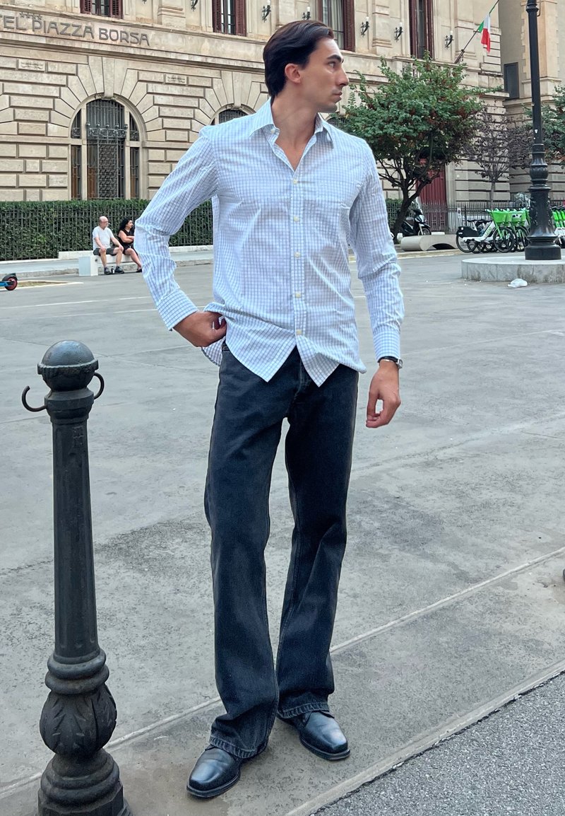 Camisa de botão branca e azul com padrão quadrado, jeans escuros e sapatos de pele pretos. De pé ao lado de um poste de iluminação em um ambiente urbano.