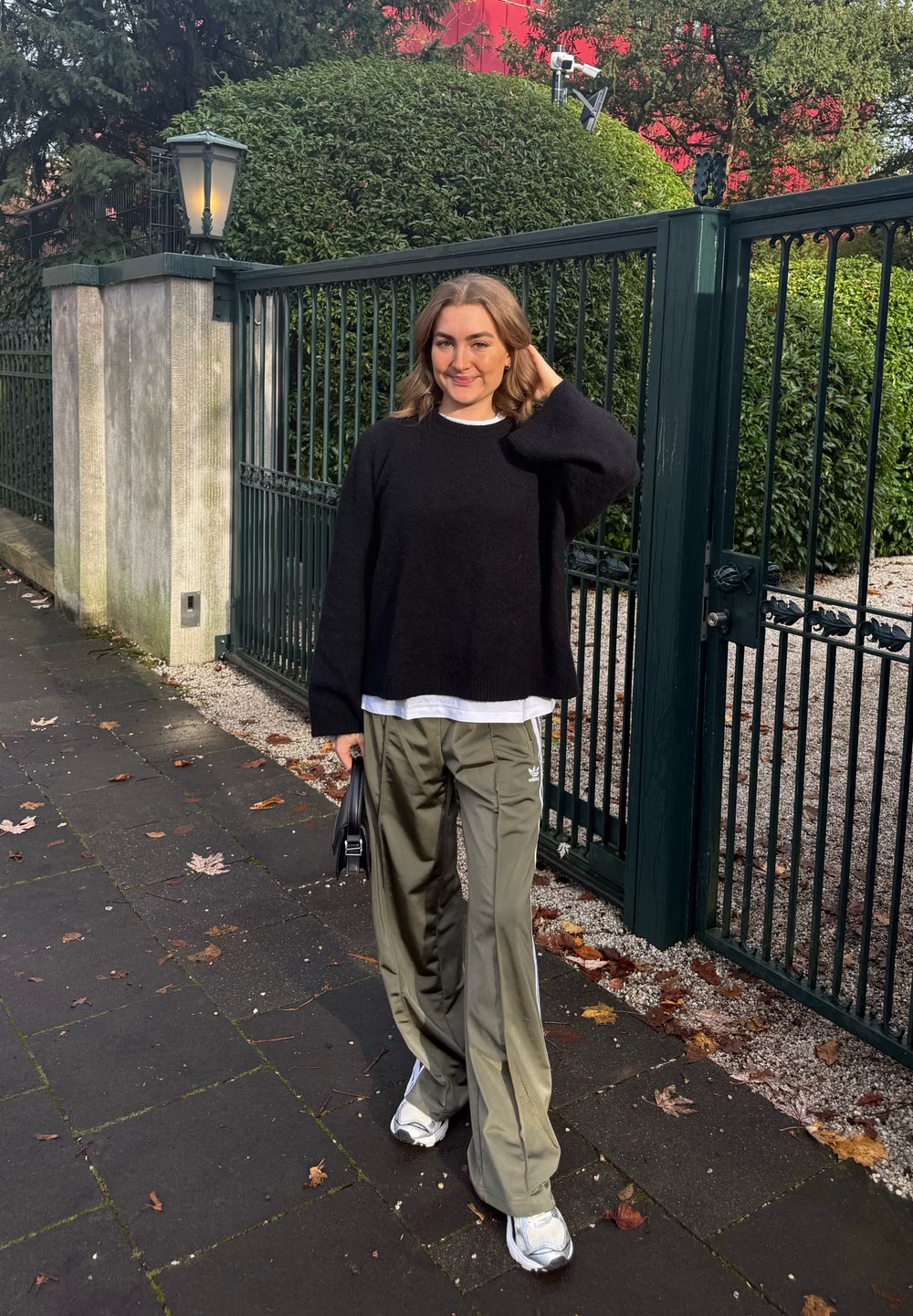 Femme en pull noir et pantalon Adidas olive tenant un sac, debout sur un trottoir près d'une clôture métallique verte et des buissons.