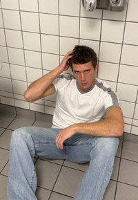 Νεαρός άνδρας με λευκή και γκρι μπλούζα και ανοιχτόχρωμα τζιν καθισμένος σε πλακάκια δάπεδο, εναντίον πλακιδίων τοίχου, με το χέρι στο κεφάλι, κοιτάζοντας μπροστά.