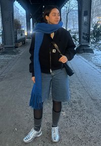 Femme debout sous un pont en vêtements d'hiver avec une écharpe bleue, une veste noire, un short en jean, des chaussettes à motifs et des baskets argentées.
