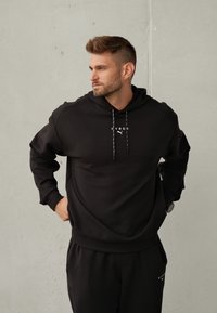 Czarna bluza z kapturem, wykonana z miękkiego materiału, z kapturem na sznurki, nadrukiem logo i ściągaczami przy rękawach. W komplecie z dopasowanymi czarnymi spodniami dresowymi.