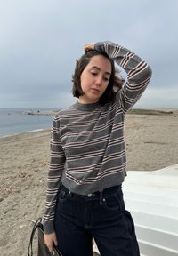 Femme aux cheveux courts et foncés, portant un pull gris rayé et un jean foncé, debout sur une plage sous un ciel couvert, tenant un sac à côté d'elle.