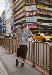 Kurzärmeliges Hemd mit schwarzem Herzmuster, weite schwarze Shorts, schwarze Sneakers und weiße Socken; urbaner Hintergrund mit bunten Schildern.