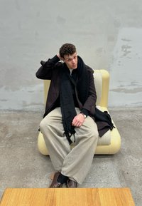 Divano curvo di un giallo chiaro con rivestimento testurizzato. Persona seduta, che indossa un lungo cappotto scuro, una sciarpa nera e pantaloni chiari. Tavolo di legno in primo piano.