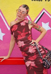 Frau mit Blumenmuster-Kleid, großer Brille, tätowiertem Arm und langen Nägeln, die sich an einem roten Geländer gegen ein lebhaftes pink-gelbes Sternenmural lehnt.