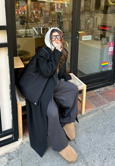 Abrigo negro de gran tamaño, pantalones de pierna ancha grises, zapatillas de ante beige, sudadera blanca con capucha, lleva unas grandes gafas de sol, sentado en un banco de madera.