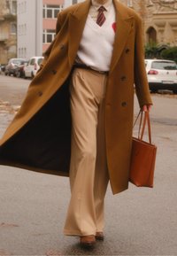 Un long manteau marron par-dessus un pull blanc avec des accents rouges, un pantalon beige et des chaussures marron ; tenant un sac à main marron en marchant dans la rue.